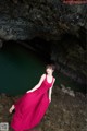 A woman in a long red dress standing in front of a cave.